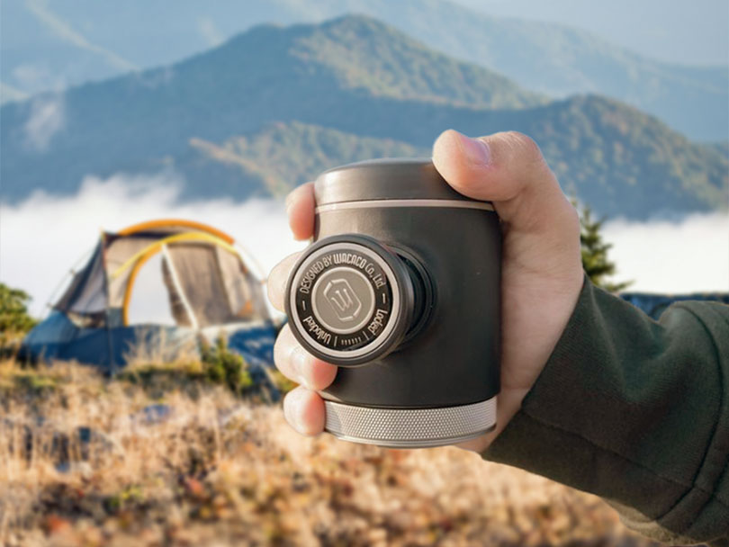 Holding the Wacaco Picopresso. portable espresso machine to the camera, tent and mountains in the background